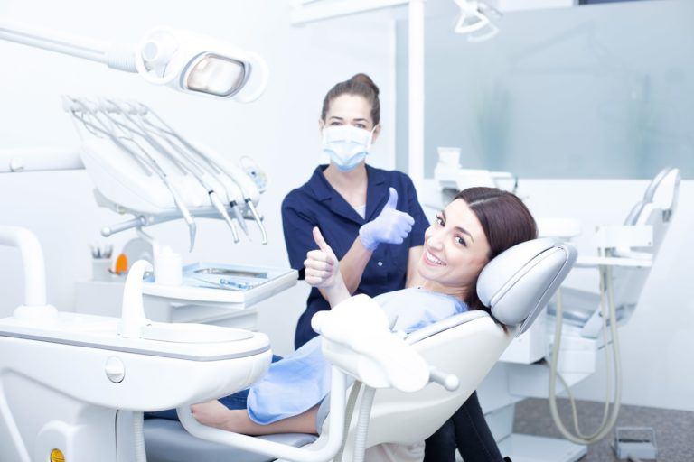 Female dentist in blue scrubs giving a thumbs up standing behind a female patient sitting in slightly reclined dental chair smiling and giving a thumbs up